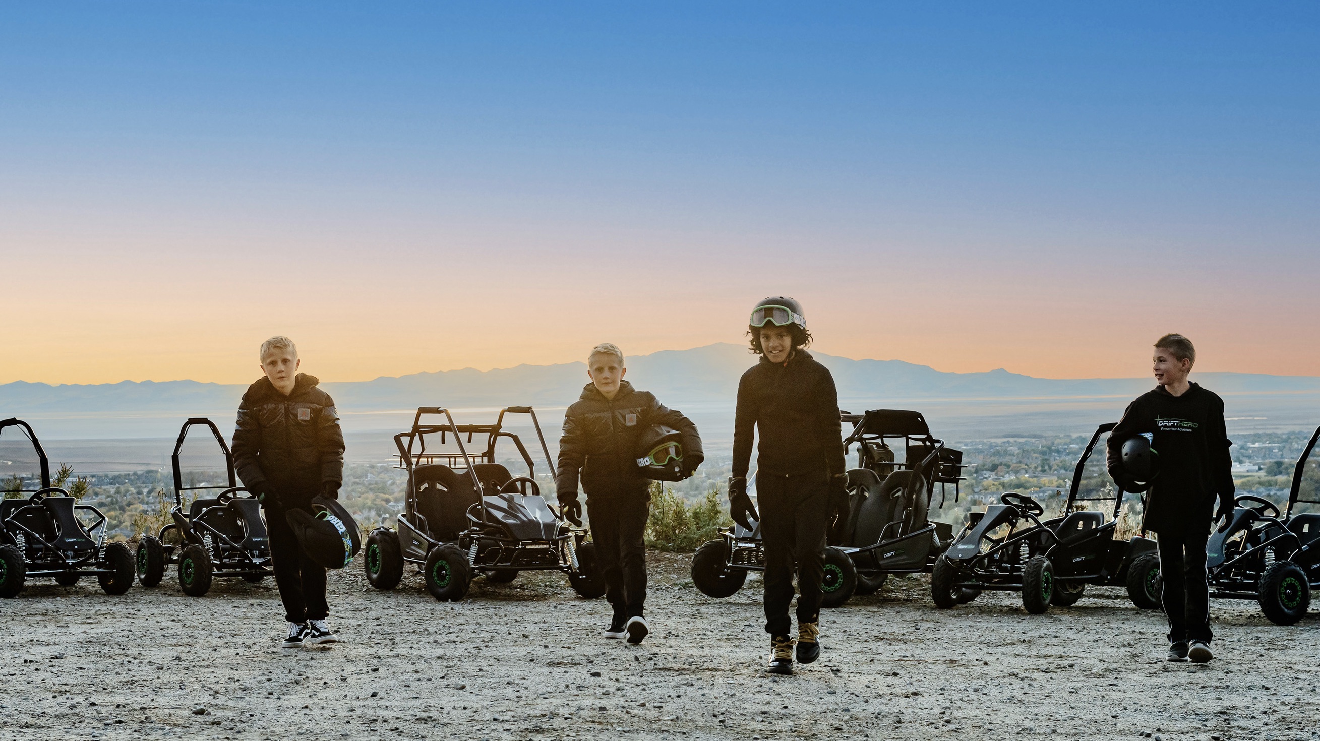 Four young riders in black riding gear walk across a gravel lot in front of a row of Drift Hero go-karts, with mountains and a sunset sky behind them